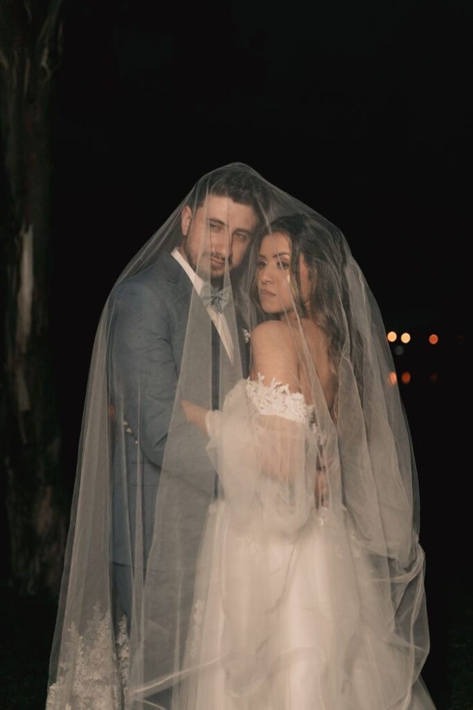 A bride and groom share a tender moment under a veil during a nighttime wedding.