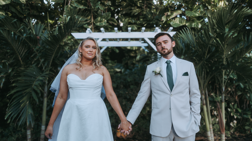 A bride and groom holding hands under a white arch in an outdoor wedding setting.